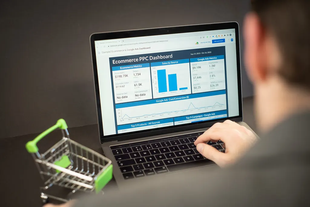 A person typing on a laptop showing an e-commerce analytics dashboard with sales, orders and ad metrics, next to a miniature shopping cart