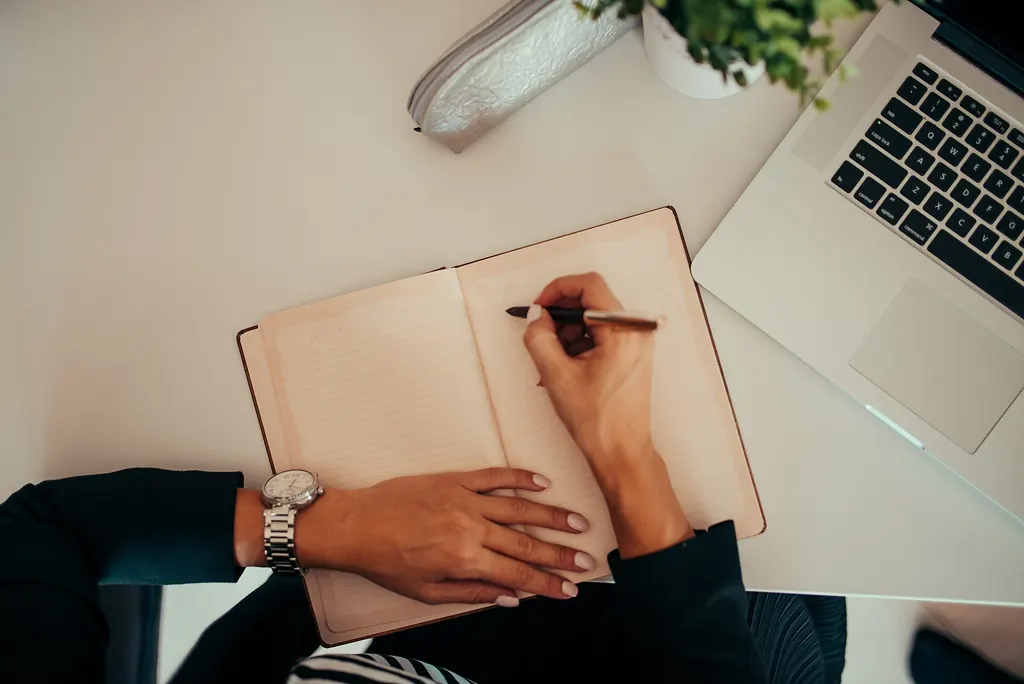 A person writing notes in a notebook next to a laptop on a white desk