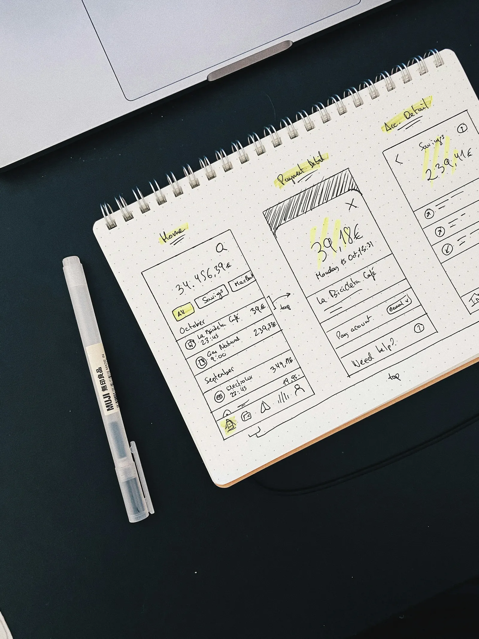 App screens sketched in a spiral notebook beside a pen